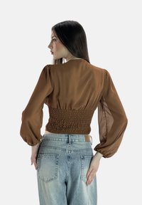 Blusa marrone trasparente con maniche a sbuffo e schiena arricciata; abbinata a jeans in denim a vita alta di colore blu chiaro.