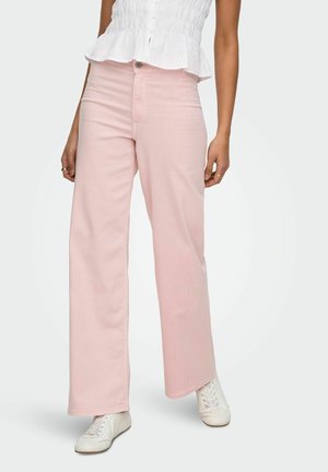 Kvinde iført lyserøde vidde bukser, hvide sneakers og en hvid ærmeløs top, stående mod en lys baggrund.