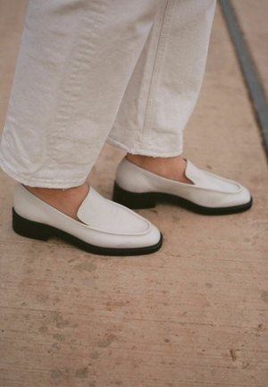 Crème leren instapschoenen met een ronde neus en een lichte zwarte hak. Draagt met witte cropped broeken op een gestructureerd oppervlak.