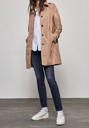 Frau trägt einen beigefarbenen Trenchcoat über einem weißen Hemd, dunkle Skinny-Jeans, weiße Socken und weiße Turnschuhe und steht auf grau gefärbtem Boden.