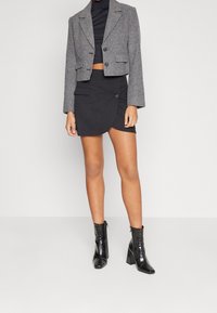 Femme portant un blazer gris court, un haut court noir, une mini-jupe noire asymétrique et des bottines noires à talons hauts, debout sur un fond uni.
