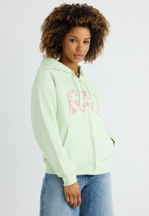 Νεαρή γυναίκα με κατσαρά μαλλιά, ντυμένη με ανοιχτό πράσινο φούτερ GAP με φερμουάρ και λουλουδάτα γράμματα, φορώντας μπλε τζιν, στέκεται μπροστά σε λευκό φόντο.