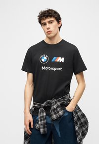 Νεαρός άνδρας με σγουρά μαλλιά φοράει μαύρο μπλουζάκι BMW Motorsport και έχει δέσει ριγέ πουκάμισο γύρω από τη μέση του, μπροστά σε απλό φόντο.