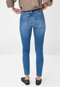 Jeans skinny en denim extensible bleu avec cinq poches, couture visible et un petit patch en cuir à la taille. Portés avec des ballerines en mesh.