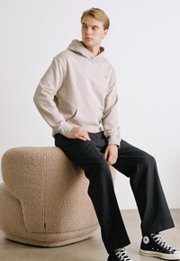 Beige kapucnis pulcsi, elülső zsebel, sötét, bő szárú nadrággal és fekete magasszárú sportcipővel viselve. Puha, textúrált ülőbútor a háttérben.