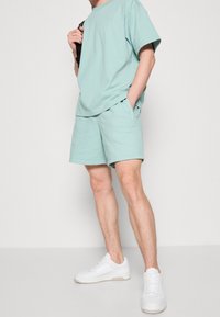 Setti mintunvihreästä puuvillapuserosta ja shortsista, jossa on rento malli, sivutaskut ja pieni logoakselti. Yhdistetty valkoisiin lenkkareihin.