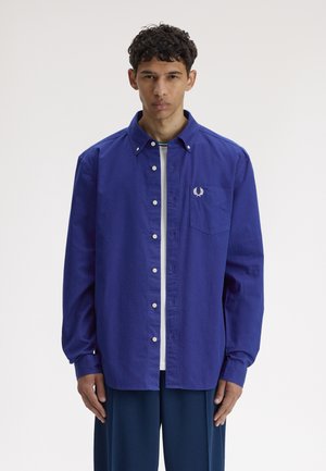 Junger Mann trägt ein hellblaues Hemd über einem weißen T-Shirt, steht vor einem schlichten weißen Hintergrund mit neutralem Gesichtsausdruck.