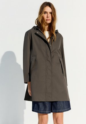 Femme portant un manteau à capuche gris foncé sur un haut blanc et un short en jean, debout devant un fond clair uni.