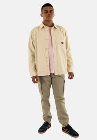 Giacca camicia in corduroy beige sopra una t-shirt rosa, abbinata a pantaloni cargo kaki chiari e sneaker scure con dettagli arancioni.