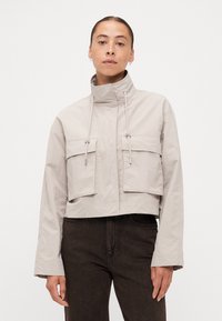 Cropped beige jacket with a high collar, adjustable drawstrings, and two large front pockets. Fabric appears smooth with a matte finish.