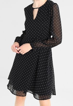 Robe noire en tissu transparent avec des manches longues, dotée d'un décolleté en V et de petits motifs géométriques blancs.