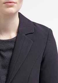 Czarna dopasowana marynarka z wyciętym kołnierzem, wykonana z gładkiego materiału z precyzyjnymi szwami, zestawiona z ciemną, wzorzystą bluzką pod spodem.