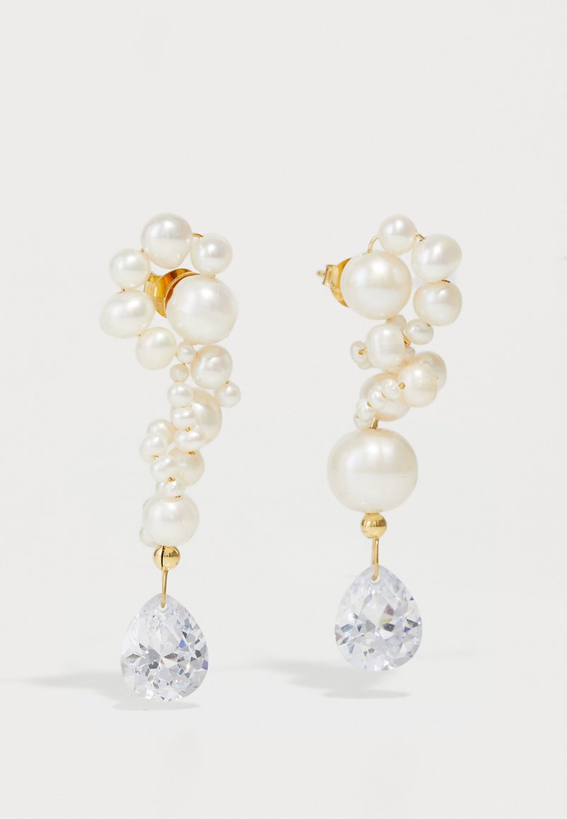 Χρυσές σκουλαρίκια με συστάδες λευκών μαργαριταριών και κρυστάλλους σε δάκρυ. Λείες και υφασμάτινες επιφάνειες δημιουργούν οπτική αντίθεση και βάθος.