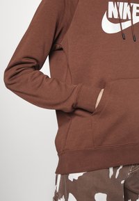 Brown sweatshirt with a large white Nike logo, featuring a kangaroo pocket and ribbed cuffs, shown with camouflage pants.