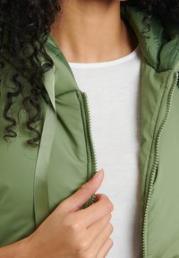 Doudoune verte avec fermeture éclair et tissu texturé. Comprend une capuche et une doublure intérieure lisse. Portée par-dessus un haut de couleur claire.