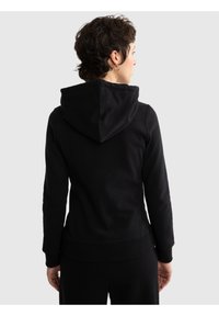 Persoană cu păr scurt și întunecat, purtând un hanorac negru, văzută din spate pe un fundal simplu și deschis la culoare.