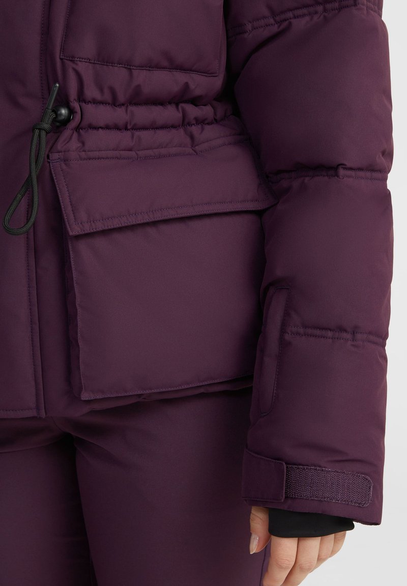 Die lila Pufferjacke verfügt über einen Kordelzug in der Taille, eine aufgesetzte Tasche auf der Brust und Klettverschlüsse an den Ärmeln.