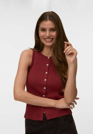Femme souriante aux longs cheveux bruns, portant un haut rouge côtelé sans manches avec des boutons et un pantalon foncé, posant avec un bras croisé et la main près du visage.
