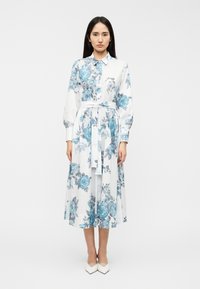 Φλοράλ φόρεμα με μπλε τριαντάφυλλα σε λευκό φόντο, με γιακά, μακριά φουσκωτά μανίκια και ζώνη με κορδέλα στη μέση.