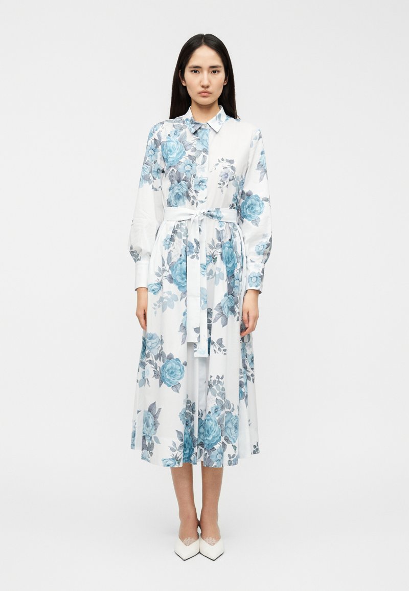 Φλοράλ φόρεμα με μπλε τριαντάφυλλα σε λευκό φόντο, με γιακά, μακριά φουσκωτά μανίκια και ζώνη με κορδέλα στη μέση.