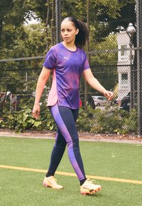 Top desportivo roxo e rosa com mangas curtas, combinado com leggings azul-marinho com um design em gradiente. Chuteiras amarelas em campo sintético.