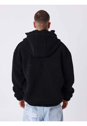 Mann trägt einen schwarzen Fleece-Hoodie mit Stern-Design auf dem Rücken, kombiniert mit hellblauen Jeans mit einem weißen Stern-Patch auf der Tasche.