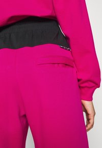 Fuchsia byxor med svart elastisk midja, med en sidoficka och slät textur. Designen betonar komfort och enkel användning.