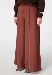 Wijde broek in roestkleur, voorzien van een riem, geplooide voorkant en gladde stof. Gecombineerd met puntige schoenen in een zwart-bruin zebra patroon.