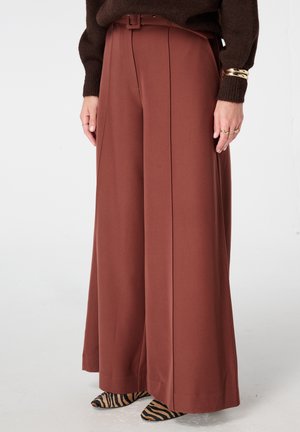 Wijde broek in roestkleur, voorzien van een riem, geplooide voorkant en gladde stof. Gecombineerd met puntige schoenen in een zwart-bruin zebra patroon.