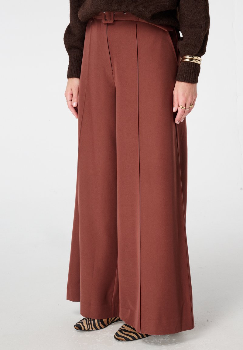 Wijde broek in roestkleur, voorzien van een riem, geplooide voorkant en gladde stof. Gecombineerd met puntige schoenen in een zwart-bruin zebra patroon.