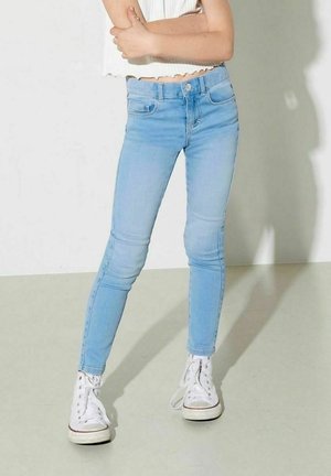 Persona que lleva vaqueros pitillo azul claro, zapatillas blancas de caña alta y una camiseta blanca corta sin mangas, de pie con los brazos cruzados.
