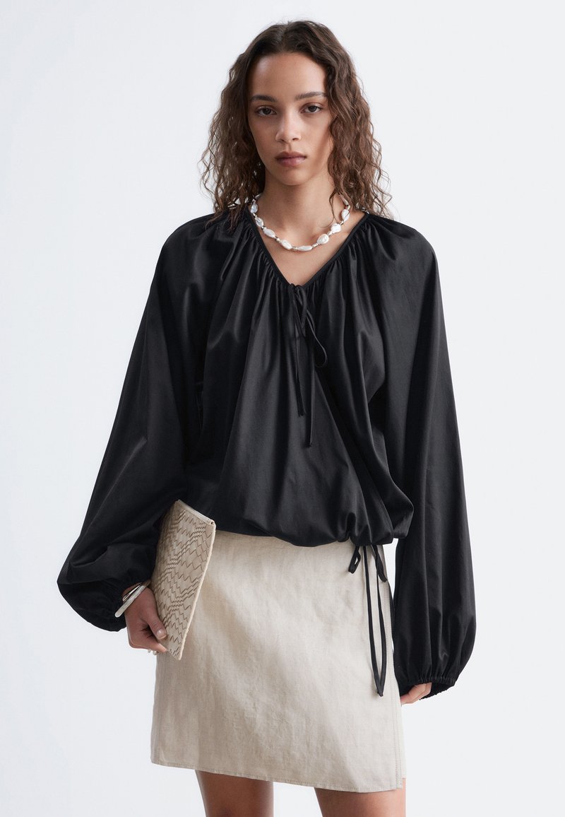 Femme aux cheveux bouclés portant un chemisier noir à manches longues et une jupe beige tenant une pochette beige texturée sur un fond uni.