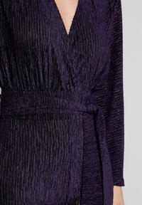 Vestido plisado púrpura con un escote en V profundo, mangas largas y un cinturón del mismo tejido. El material tiene un efecto de brillo texturizado.