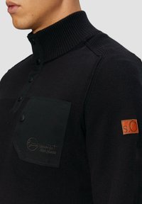 Dunkler Strickpullover mit hohem geripptem Kragen, schwarzer aufgesetzter Tasche, schwarzen Knöpfen und einem braunen Leder-Label auf dem rechten Ärmel.