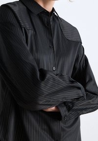 Black button-up shirt with thin vertical stripes in dark gold, featuring a collar, long sleeves, and a relaxed fit.