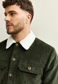 Chaqueta de corduroy verde oliva con un cuello de sherpa blanco, que presenta dos bolsillos en el pecho y botones de latón. Tejido texturizado con costillas verticales.