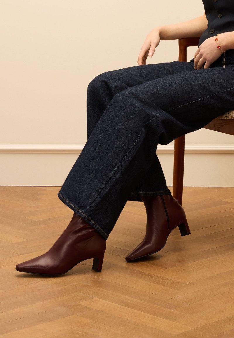 Bruine leren enkellaarsjes met een spitse neus en blokhak, gecombineerd met donkere denim wijde broek, zittend op een houten stoel.