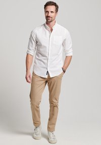 Hvid skjorte med knapper og en frontlomme, parret med beige bukser og hvide sneakers. Glat tekstur med opsmøgede ærmer og enkelt design.