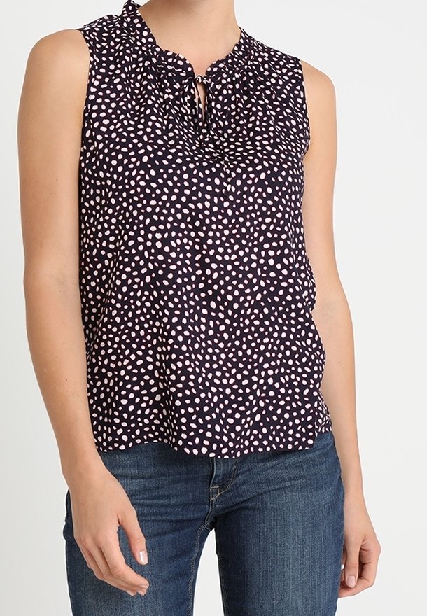 Černá bezrukávová blůza s růžovými puntíky a vázáním kolem výstřihu, nošená s modrými džínami.