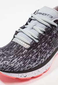 Gros plan d'une chaussure de course Under Armour noire et grise à motifs, avec des lacets gris clair et une semelle blanche à taches noires et une semelle extérieure rose.