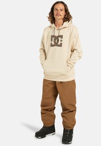 Beżowy hoodie z dużym brązowym logo, kieszenią na froncie i sznurkiem ściągającym; zestawiony z brązowymi wodoodpornymi spodniami i czarnymi śniegowcami.