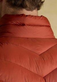 Červená prešívaná bunda s vysokým golierom, hladkou textúrou a jemn�ým logo vzorom na zadnej strane. Zdobí ju prešívanie v tvare V.