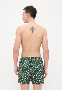 Grønne svømmebukser med et hvidt abstrakt mønster, elastisk talje og en baglomme. Stoffet fremstår let og glat.