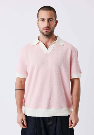 Jonge man met kort haar en baard, die een roze-wit gestructureerd polohemd met korte mouwen en een donkere broek draagt, staande tegen een effen achtergrond.