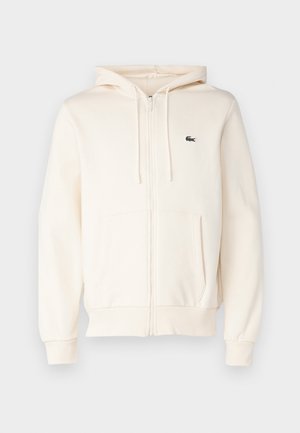 Kremfarget zip-up hettegenser med kengurulomme, snorhette, ribbestrikkede ermer og en liten mørk logo på venstre bryst. Mykt tekstilmateriale.