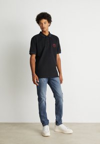 Camisa polo negra de manga corta, con un logo circular rojo en el pecho, combinada con jeans de mezclilla azul claro y zapatillas blancas.