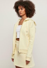 Sudadera amarilla con cremallera, capucha y bolsillos frontales, combinada con una falda de cintura elástica a juego. La tela parece suave con una textura lisa.
