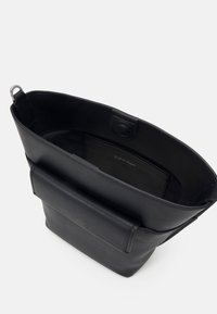 Sac seau en cuir noir avec une poche à l’avant, une fermeture magnétique au sommet et une étiquette Calvin Klein visible à l’intérieur.