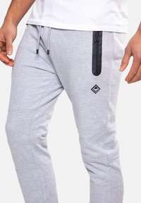Graue Jogginghose mit einer schwarzen Reißverschlusstasche, schmaler Schnitt, elastischem Bund und dekorativen Nähte an den Beinen.