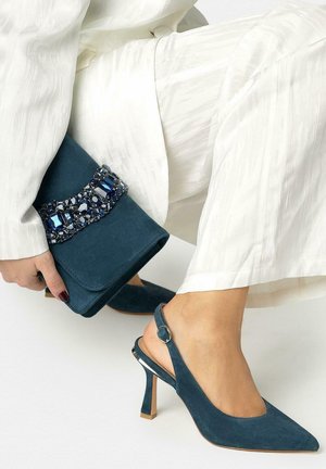 Femme portant un pantalon blanc tenant une pochette en daim bleu ornée de pierres précieuses tout en montrant des escarpins slingback en daim bleu assortis.